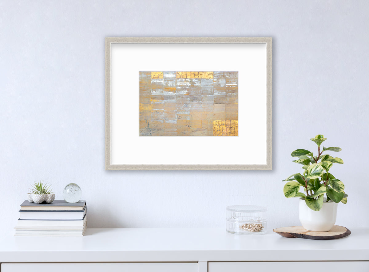 A grey, silver and gold abstract geometric print in a silver frame with a mat hangs on a white wall above a white surface with a stack of books, a round clear container with gold accessories and a potted plant.
