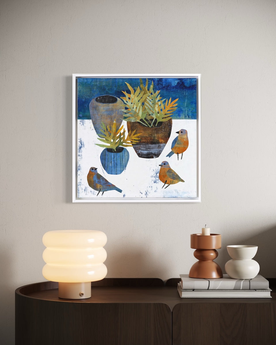 A framed collage featuring blue birds and potted plants, displayed on a light gray wall above a dark wood sideboard with a glowing lamp and decorative objects.