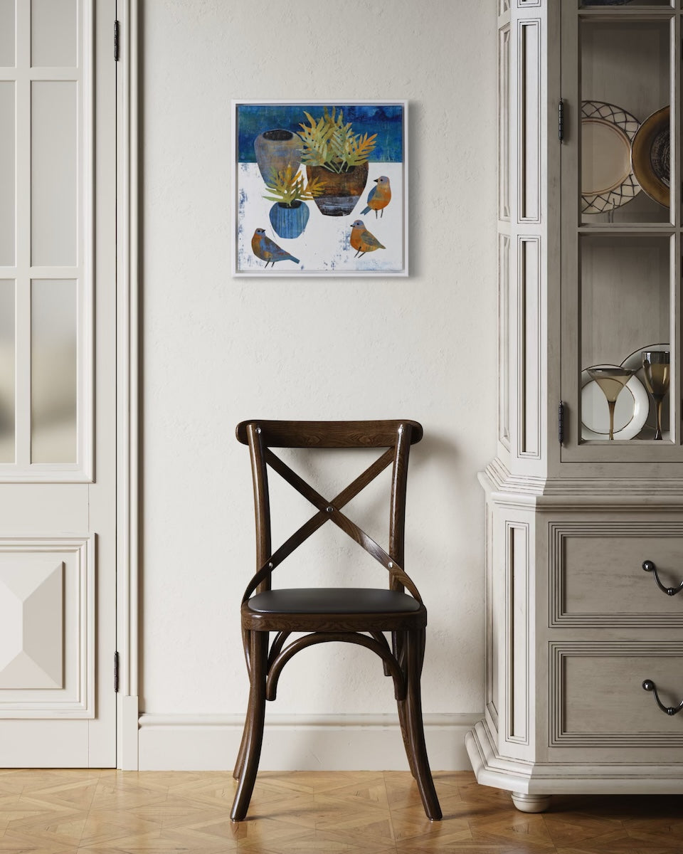 A framed square collage painting depicting potted plants and birds, displayed on a cream wall above a dark wooden chair in a styled room with a cabinet and herringbone wood floor.