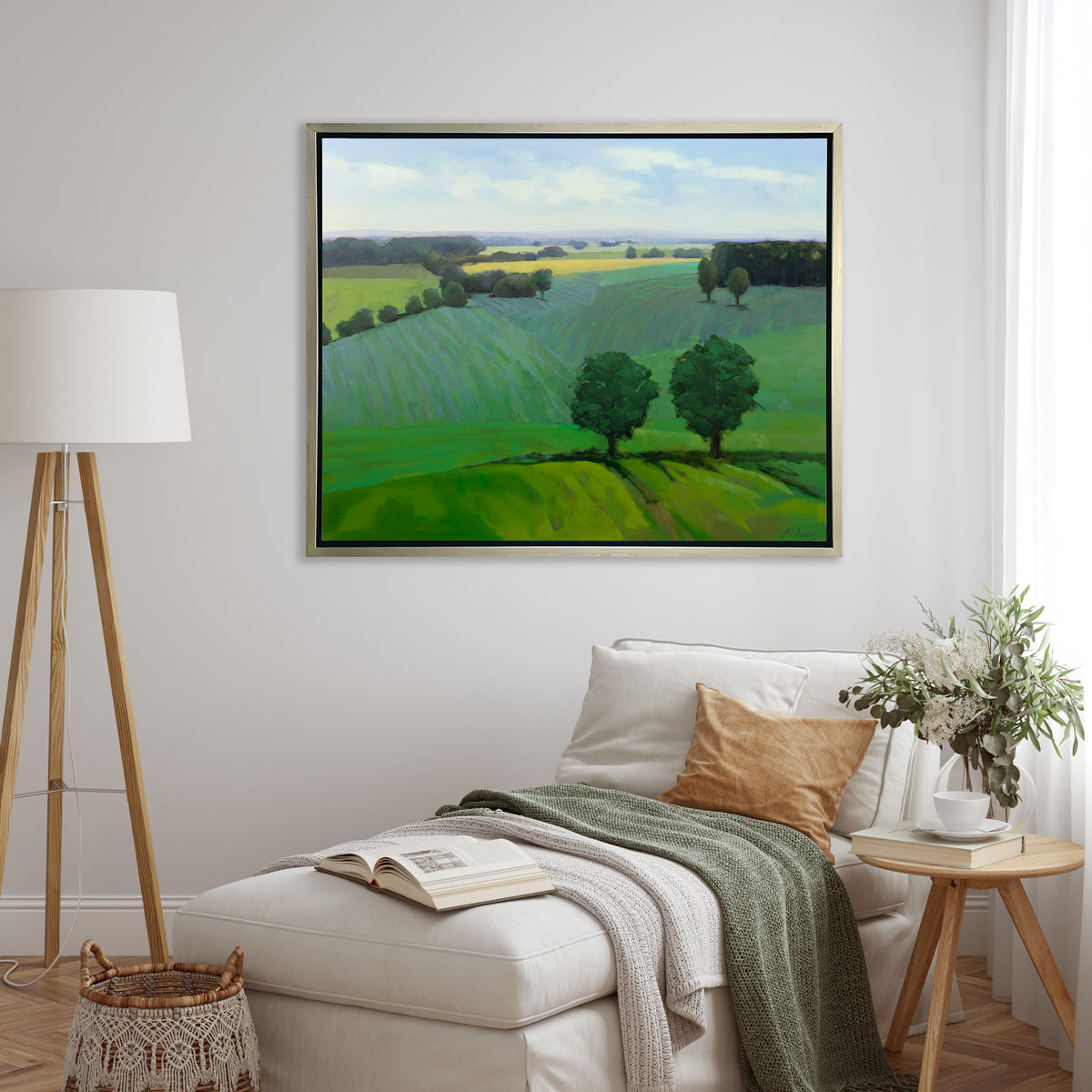A landscape print in a silver floater frame hangs on a white wall above a white chaise lounge with neutral pillows, a green and white throw blanket, a small side table with decorative accessories and a wooden tripod floor lamp.