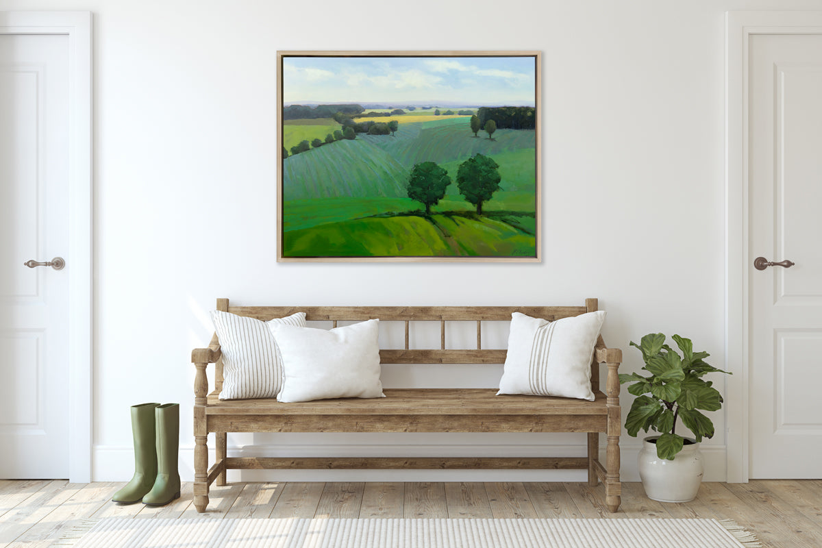 A landscape print in a white oak wood floater frame hangs on a white wall above a natural wood bench with neutral pillows, a small potted plant and a pair of green rubber boots.