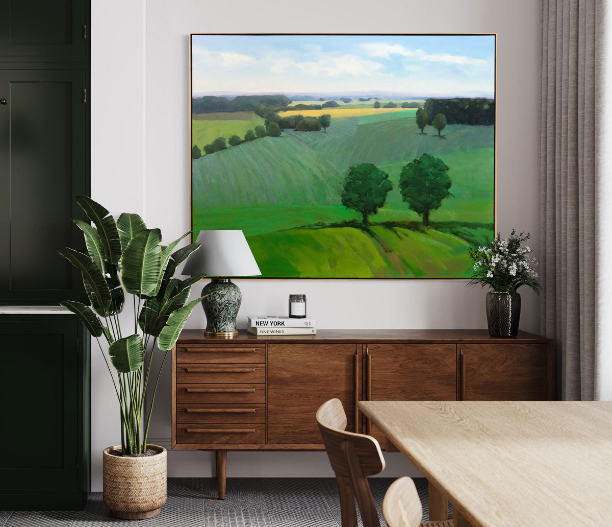A large green landscape painting hangs on a white wall above a wood console table with table decor and a floor plant. 