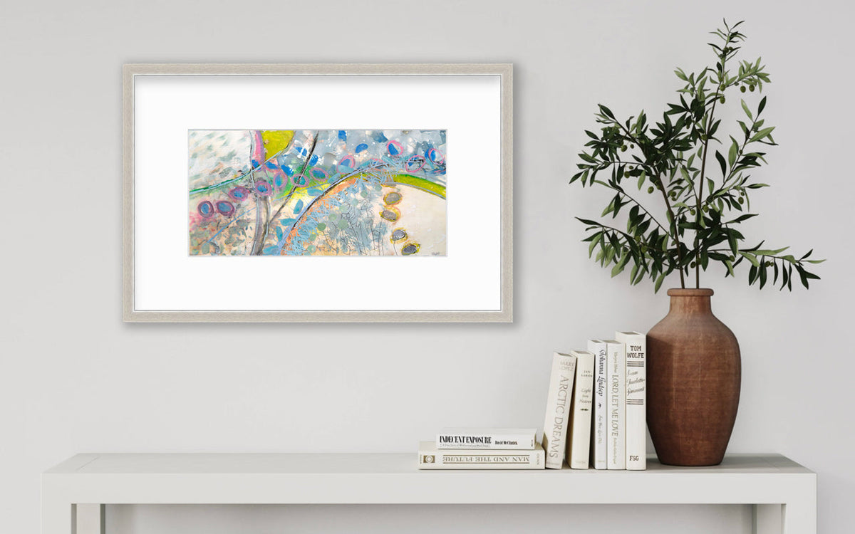 A multicolored abstract print in a silver frame with a mat hangs on a white wall above a white side table with a stack of books and a terracotta vase with greenery stems.