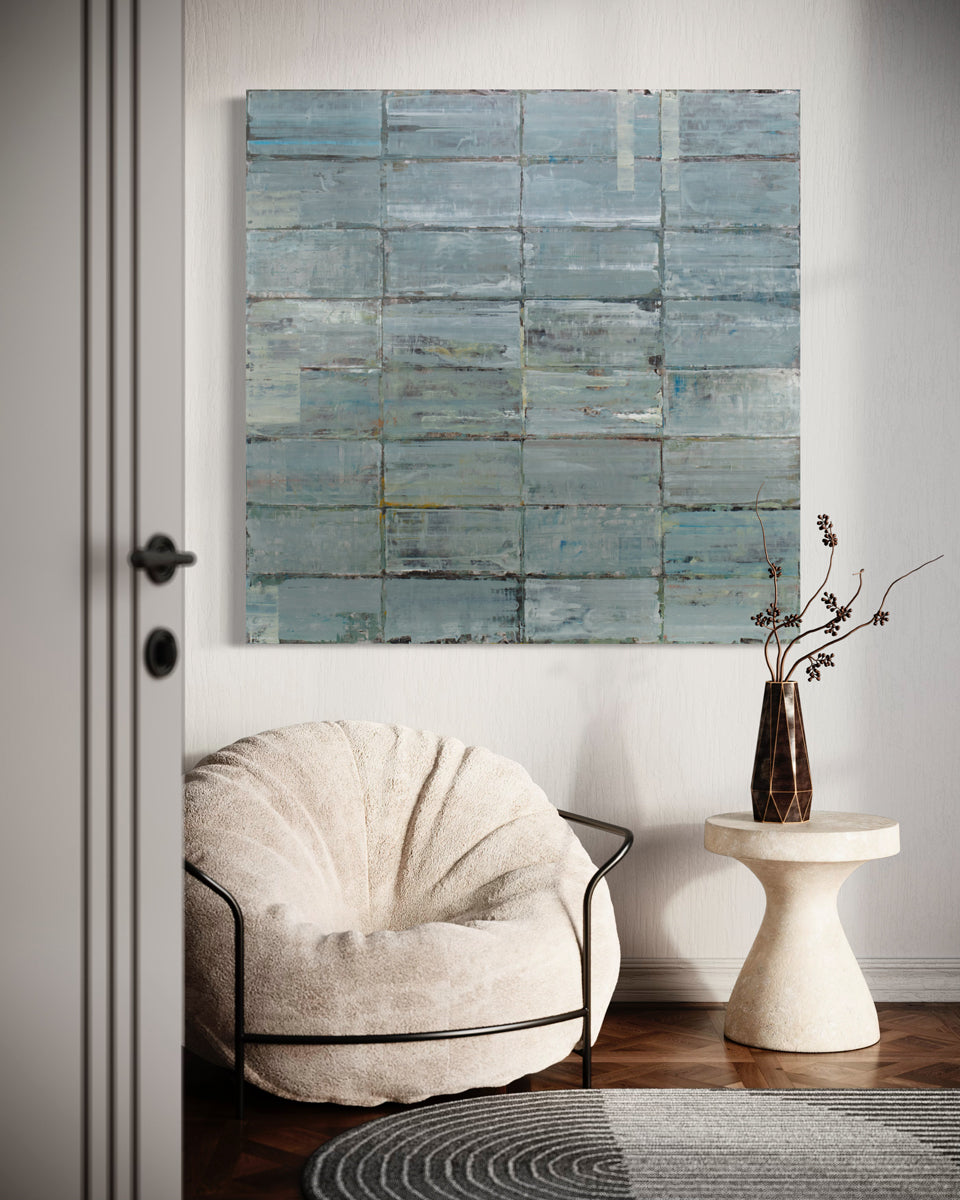 A blue and grey abstract geometric painting hangs on a white wall above a modern armchair and end table. 