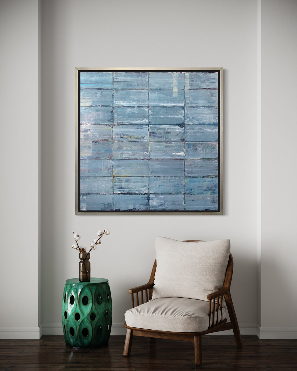 An abstract blue-green limited edition art print by Ned Martin framed in a silver frame hangs on a white wall with a brown and white armchair and a green side table.