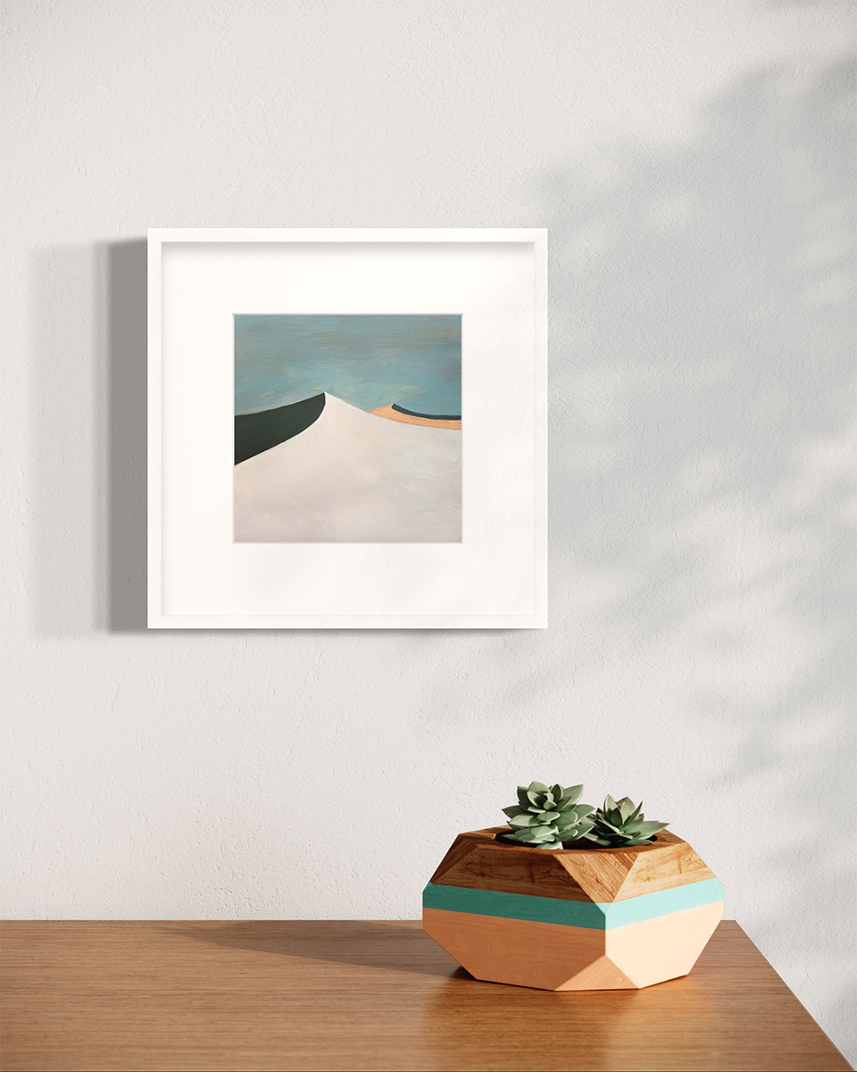 A blue, white, peach and dark teal abstract landscape print in a white frame with a mat hangs on a white wall above a wood surface with a succulent arrangement in a wood planter.