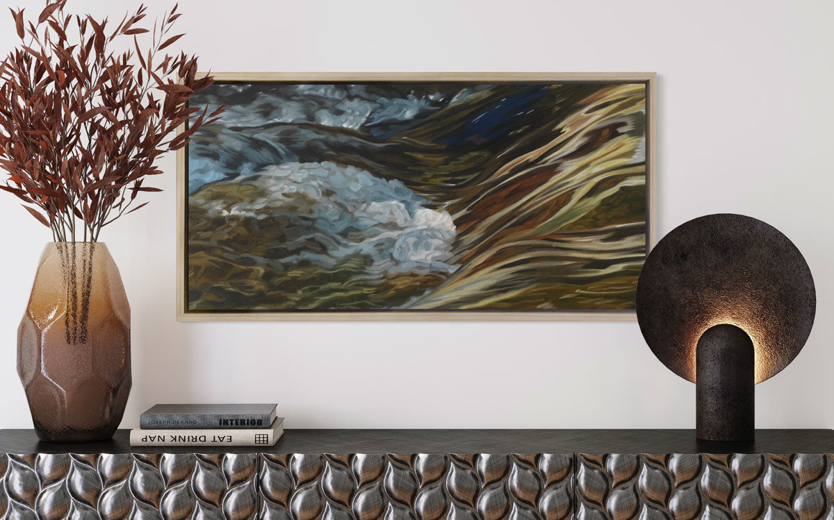 A painting of bubbling water flowing in a rocky river in a white oak frame hangs on a white wall over a carved silver mantel with a vase, books and other decor.