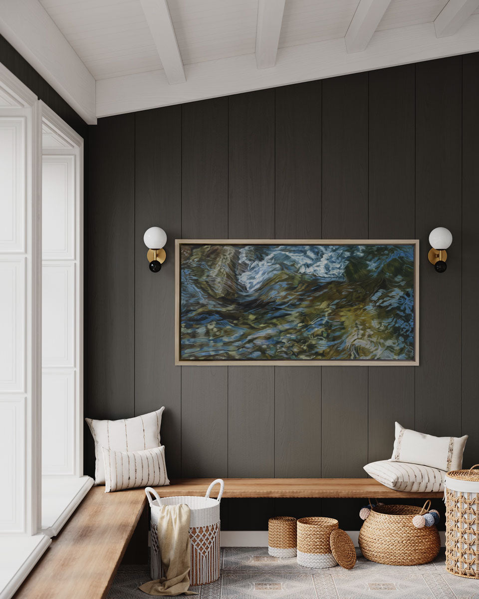 A painting of flowing water in a rocky riverbed in a white oak frame hangs on a brown wall over a wooden-built in bench with cream-colored pillows and woven baskets.