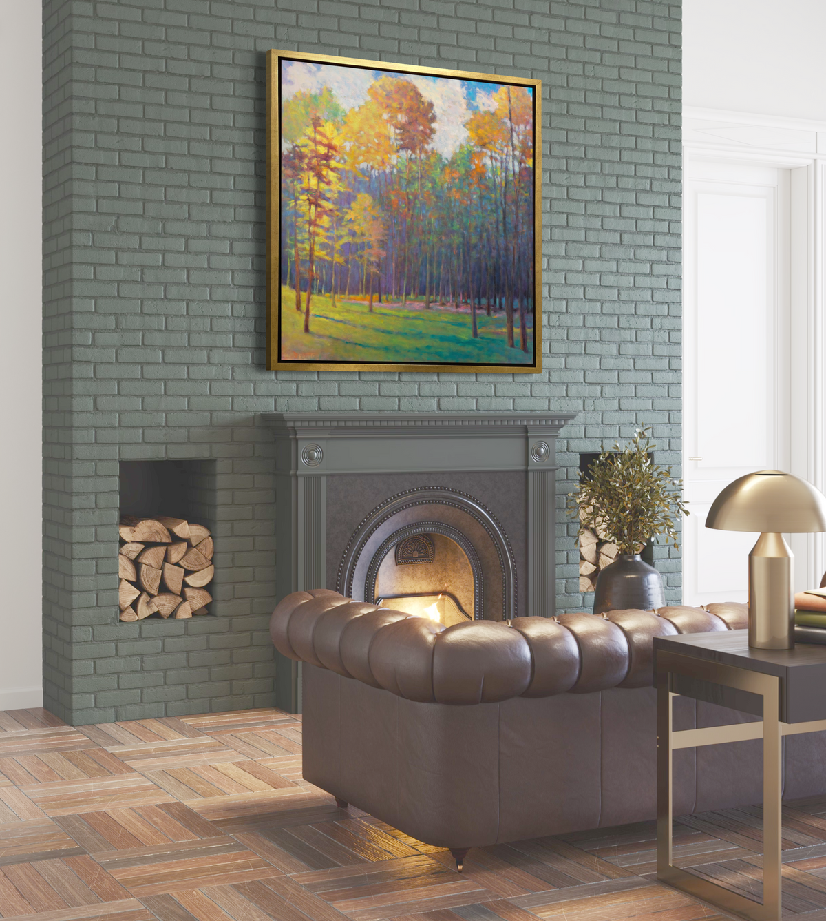 A side angle view of an impressionist landscape print in a gold floater frame hangs on a soft green brick chimney above a lit fireplace and an overstuffed leather sofa.
