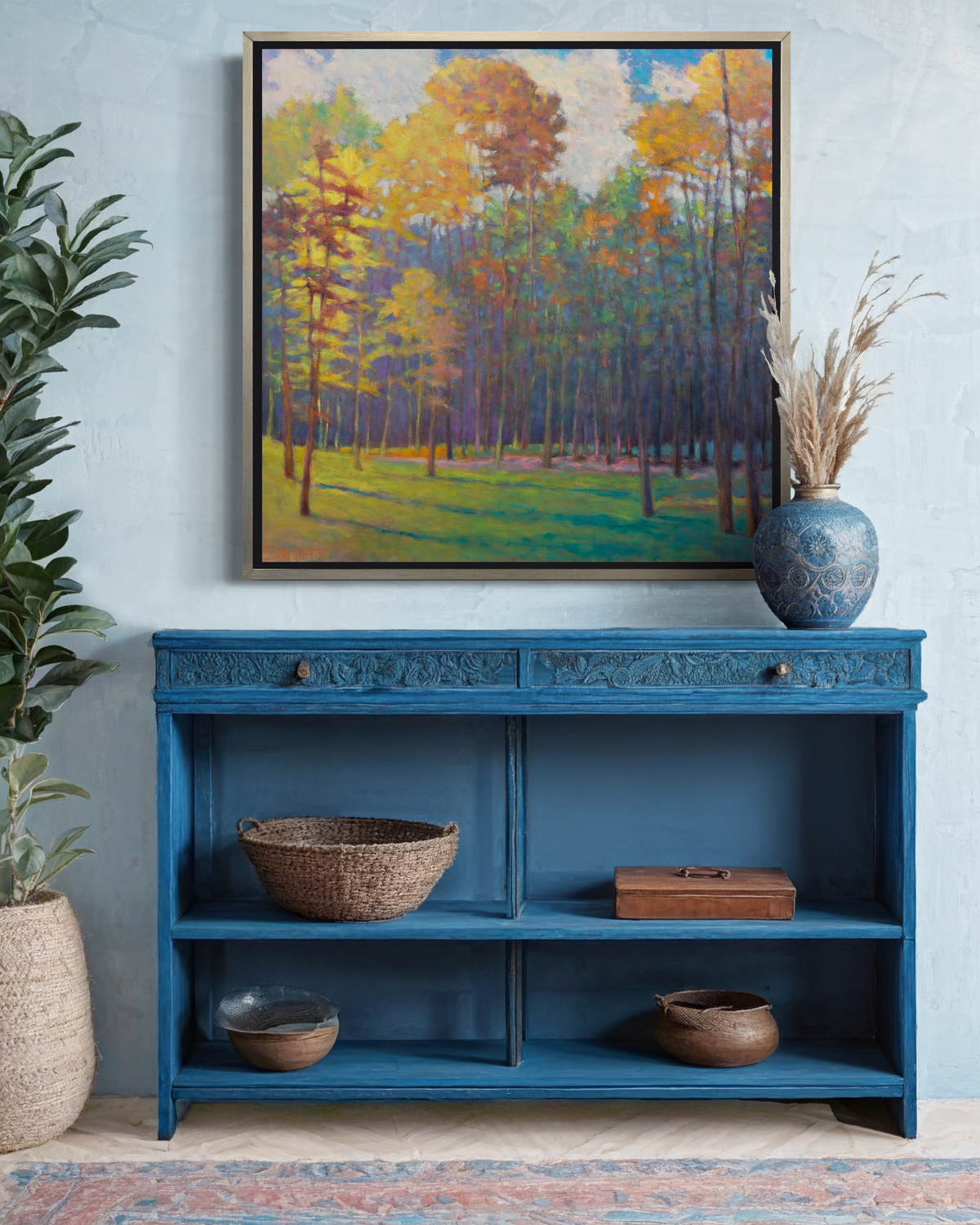 An impressionist landscape print in a silver floater frame hangs on a white wall above a blue open cabinet with shelves for baskets and boxes and a blue vase sits on top.