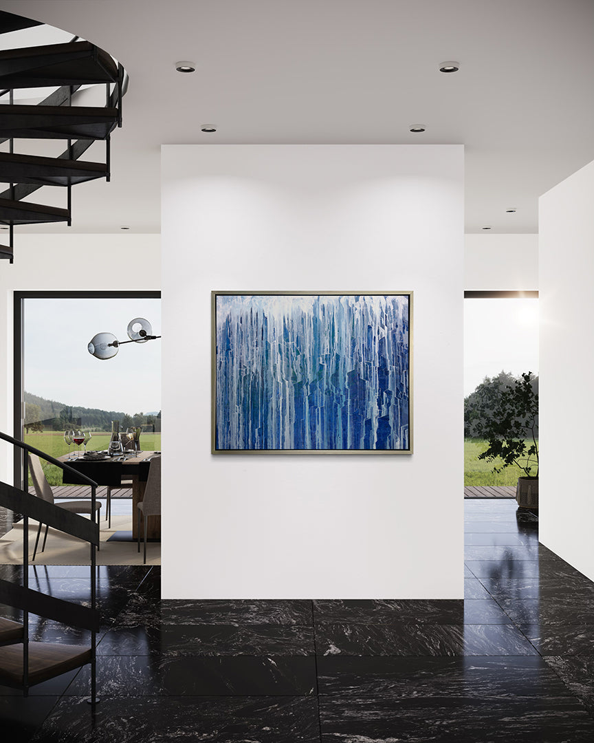 A blue and white abstract print in a silver floater frame hangs on a white wall in a foyer with a circular staircase to the left.  The kitchen is seen behind this wall with large windows to the yard beyond.