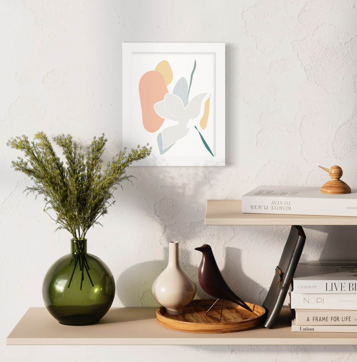 A light abstract botanical art print in an unmatted white frame hangs on a white textured wall above two natural wood floating shelves with stacks of books, a green vase with greenery and decorative accessories.