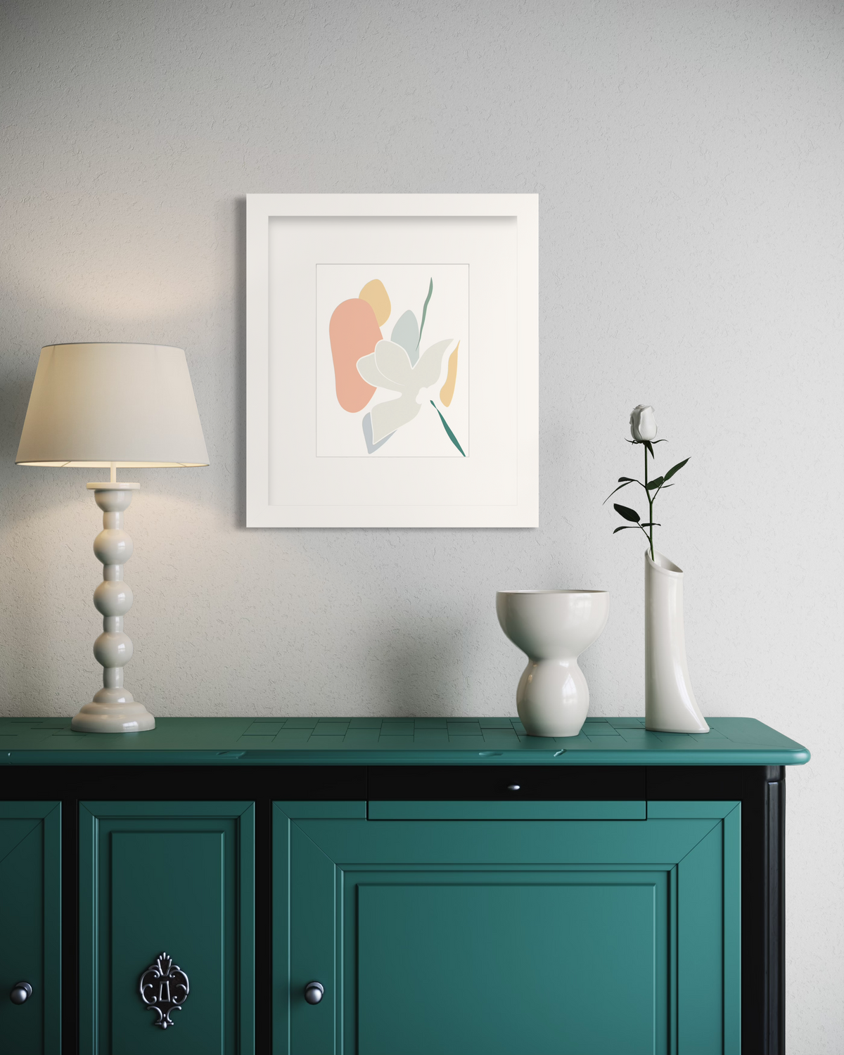 A light abstract botanical art print in a white frame with a mat hangs on a white wall above a teal cabinet with a white table lamp, a short white decoration and a white vase with a white rose.
