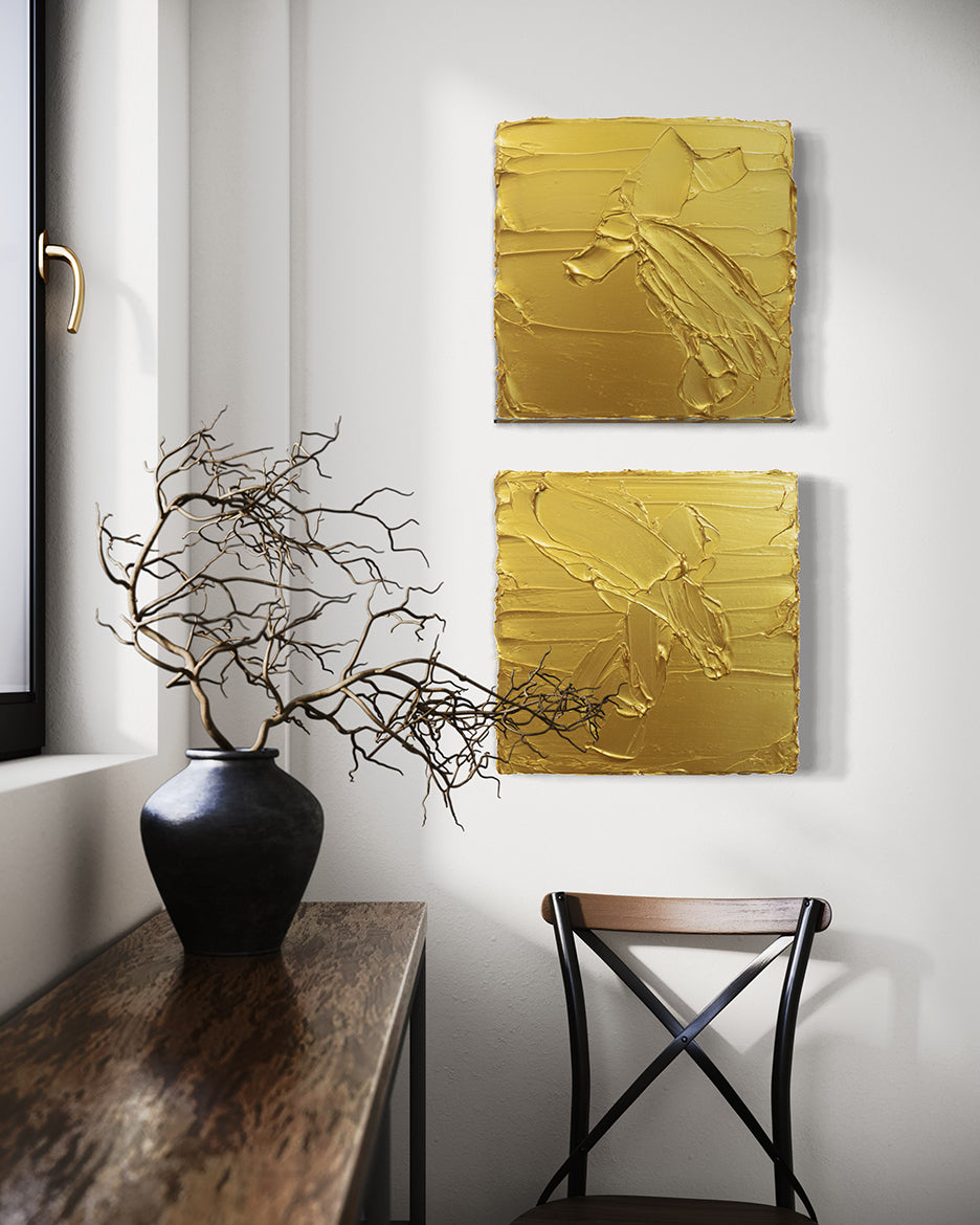 A pair of golden textured paintings by Teodora Guererra hanging on a wall above a black and brown chair. To the left is a wood table under a window with a black ceramic vase holding dried branches.