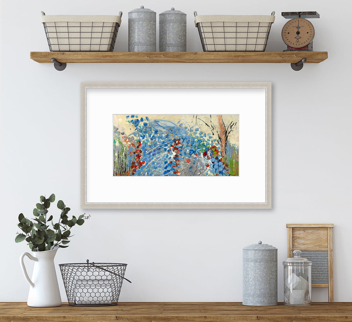 A blue, beige, green and black abstract print in a silver frame with a mat hangs on a white wall below a floating wood shelf with wire baskets and grey canisters and above a wood surface with a white pitcher holding eucalyptus stems, a wire basket and decorative accents.