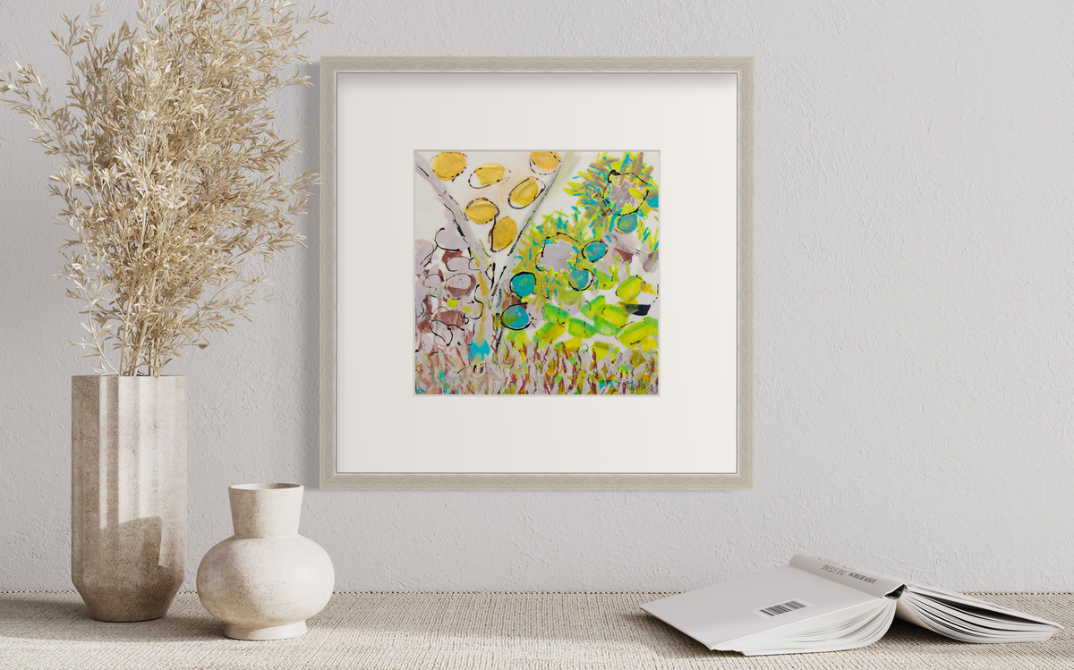 A yellow, green and red abstract floral print in a silver frame with a mat hangs on a white wall above a cloth surface with an open book, a short white vase and a taller white vase with wheat stems.