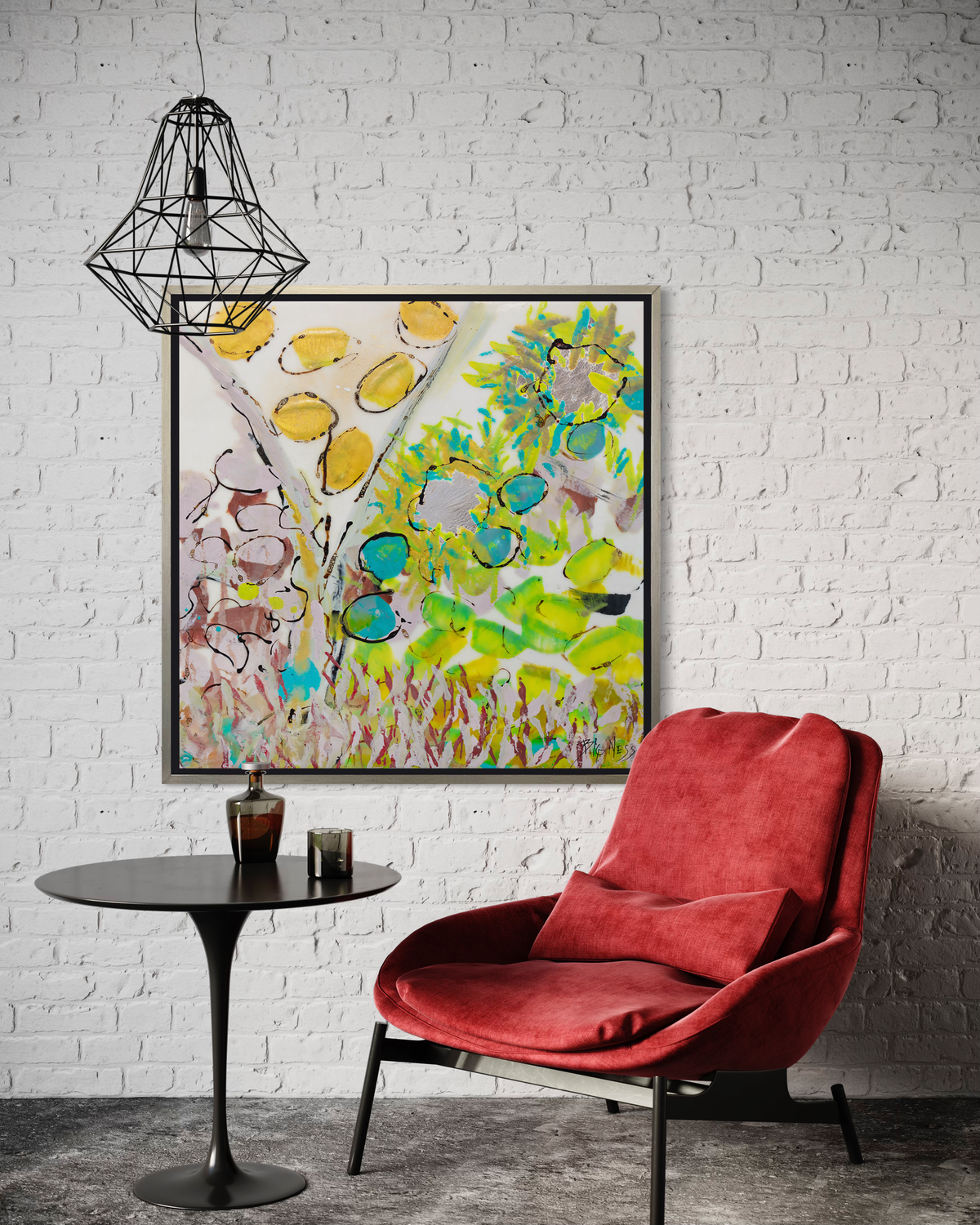 A yellow, green and red abstract floral print in a silver floater frame hangs on a white brick wall above a low red chair and a black pedestal table with drinking glasses.
