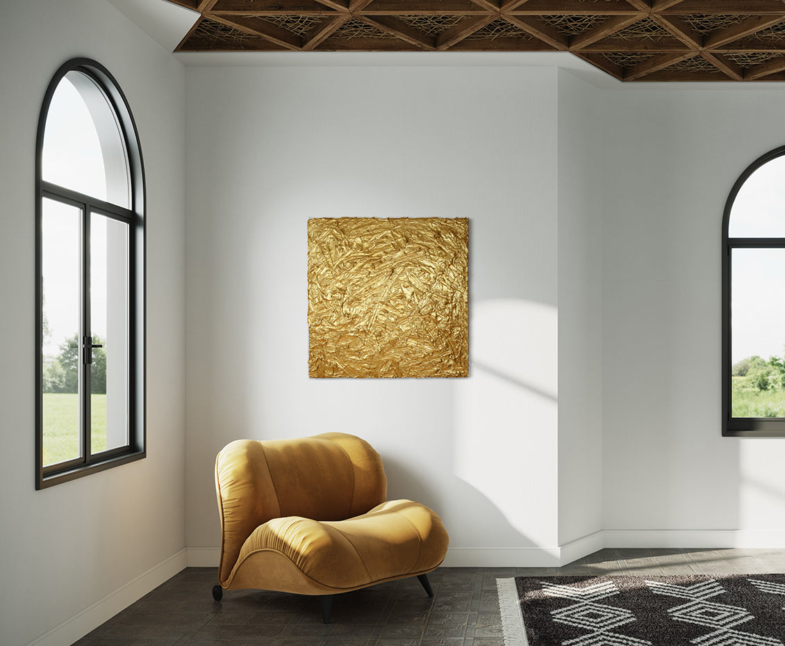 A metallic gold thick textured painting on a white wall in natural light by Teodora Guererra hangs over a yellow ochre velvet modern chair.  To the left and right are windows and in front is a carpet in a black and white pattern.