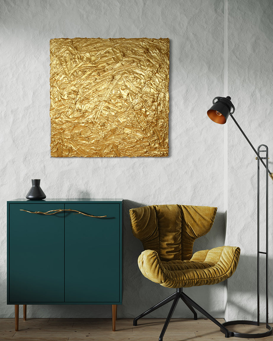 A metallic gold thick textured painting on a white wall in natural light by Teodora Guererra hangs over a green cabinet with brass tree branch like handles and a black ceramic vase sits on top.  To the right is a yellow ochre velvet modern chair and a black floor lamp.
