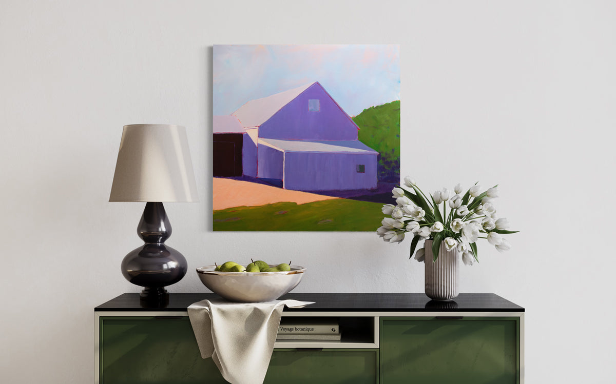 A landscape painting of a barn in lavender shadow, blue sky and green hedges by Carol Young hangs on a white wall above a black and deep green credenza with a bowl of pears, a white vase filled with white tulips and a black modern lamp table lamp.
