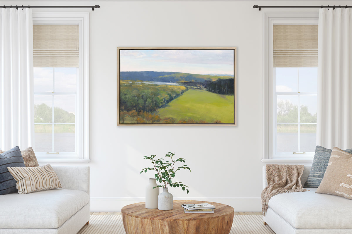 A landscape print in a white oak wood floater frame hangs on a white wall in between two windows, two white sofas with neutral pillows and a solid round wood coffee table.