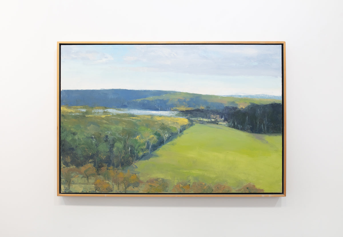 A yellow-green landscape painting in a light wood frame hangs on a light wall.