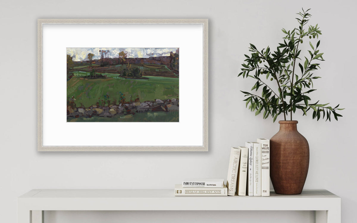 A green, brown and white abstract landscape print in a silver frame with a mat hangs on white wall above a white side table with a stack of books and a terracotta vase with greenery stems.