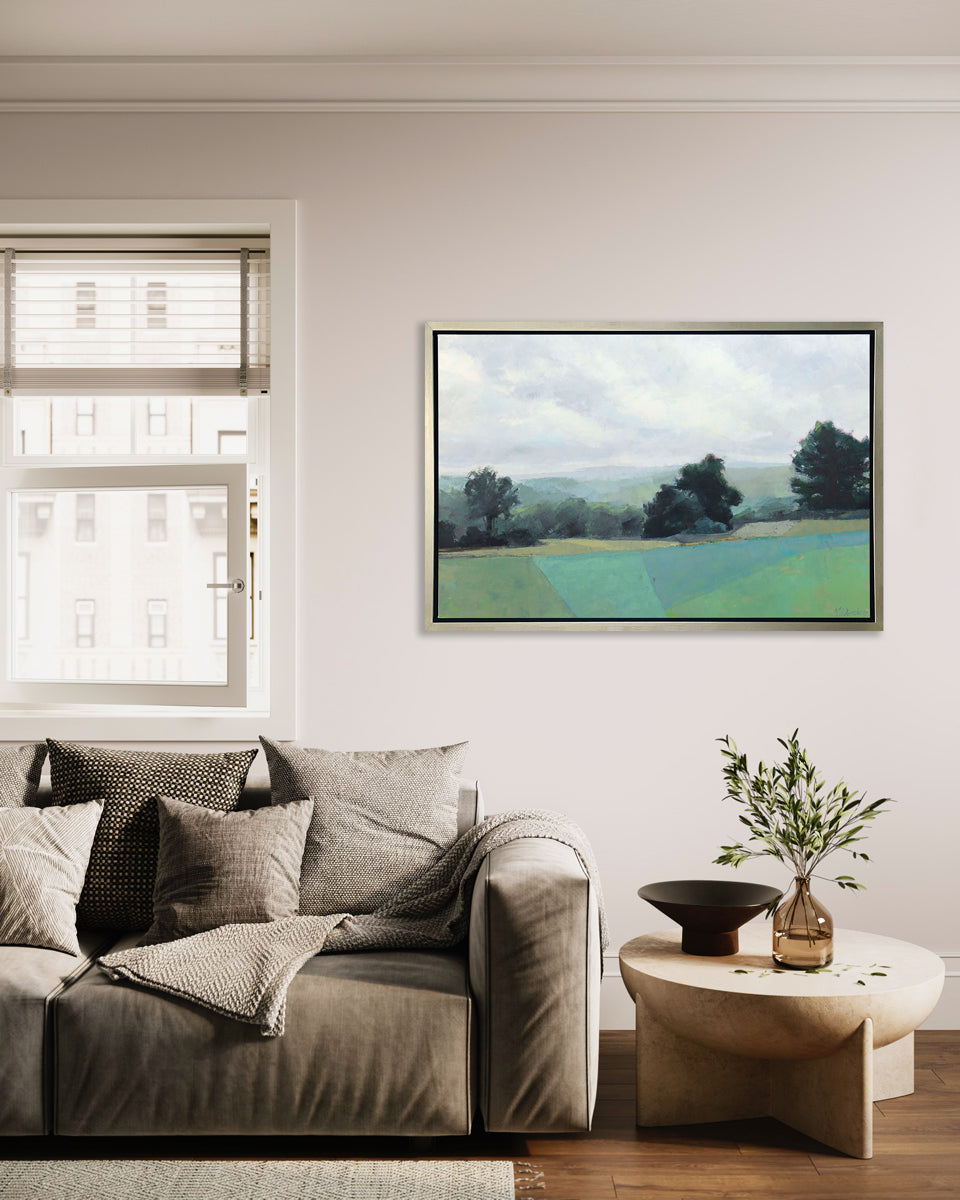 A landscape print in a silver floater frame hangs on a white wall above a grey sofa with similar colored pillows, a throw blanket and a short rounded side table with decorative accessories.