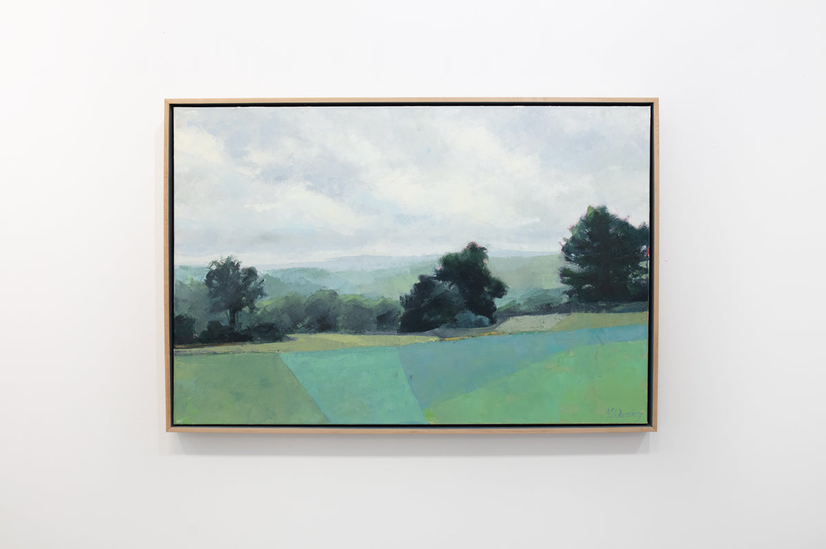 A green landscape painting framed in a light wood frame hangs on a white wall. 