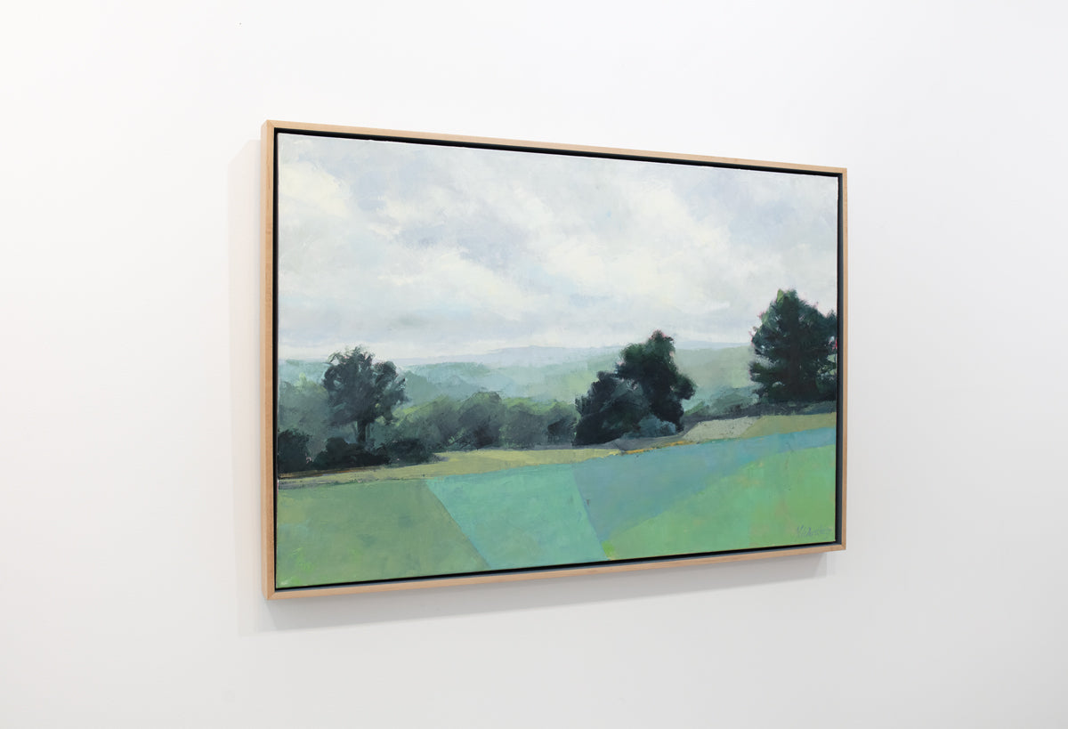 A green landscape painting framed in a light wood frame hangs on a white wall.