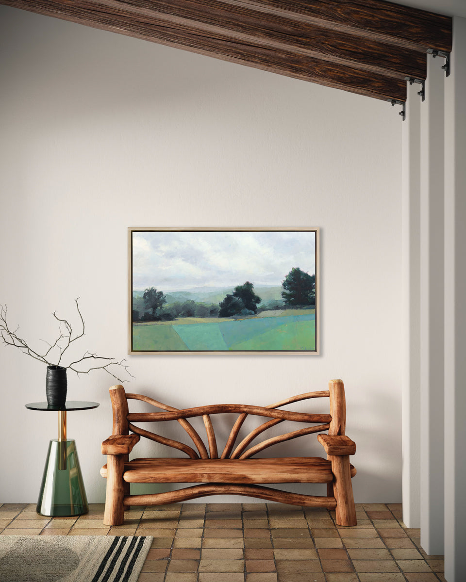 A landscape print in a white oak wood floater frame hangs on a white wall above a carved wood bench and a modern green glass side table with a black vase holding branch stems.