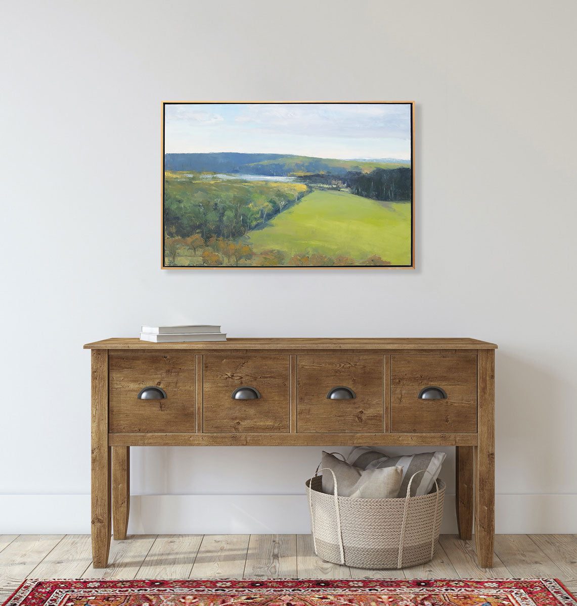 A yellow-green landscape painting in a light wood frame hangs on a light wall above a wood console table. 