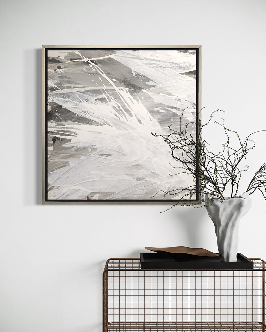 A grey, beige and white abstract print in a silver floater frame hangs on a grey wall above a wire shelf with a white ceramic vas holding decorative branches.