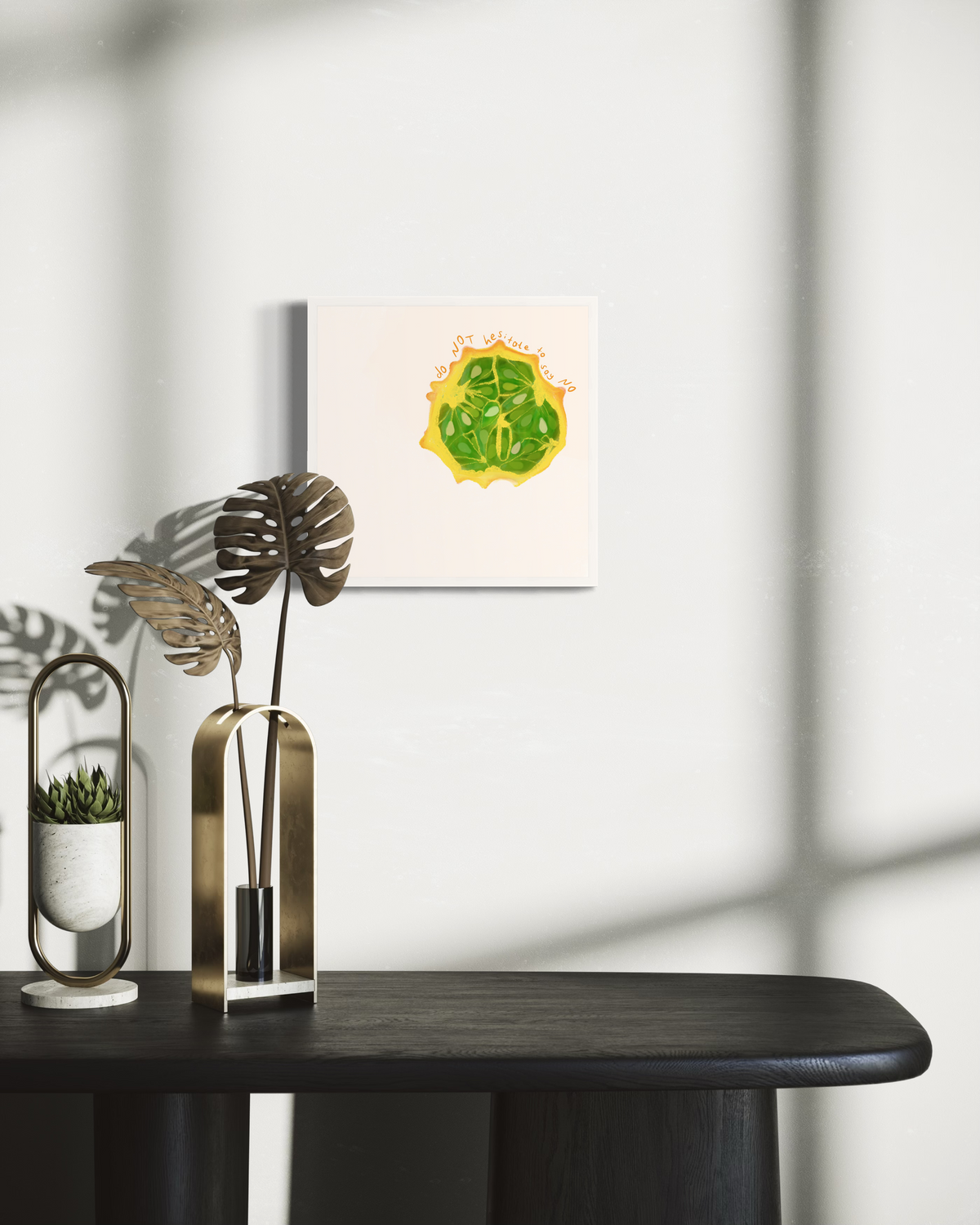 An art print of a fruit with the words "do not hesitate to say no" in an unmatted white frame hangs on a wall above a black table with two flora decorations. 
