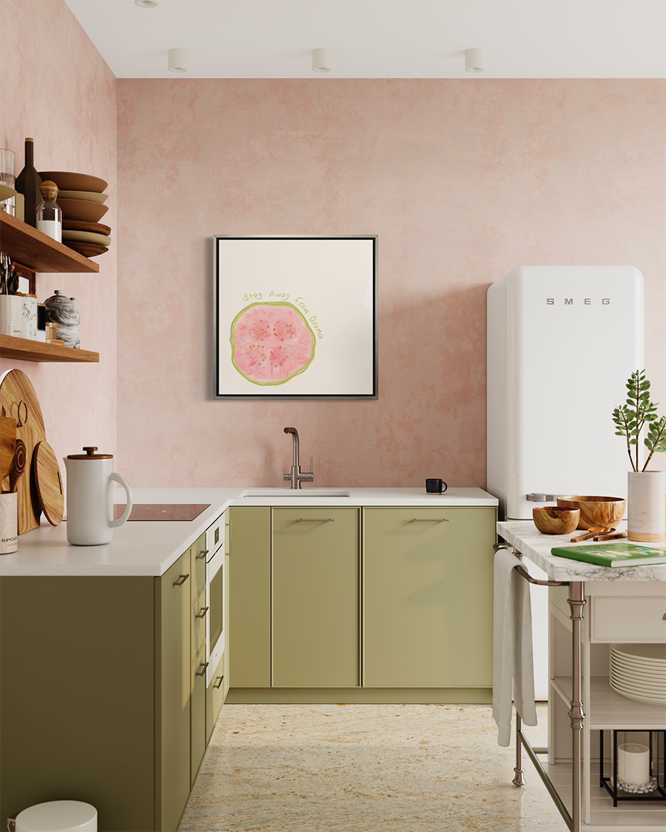 An abstract art print of a fruit framed in a silver floater frame hangs on a light pink wall above light green kitchen cabinets, a white vintage narrow refrigerator and a white marble island.