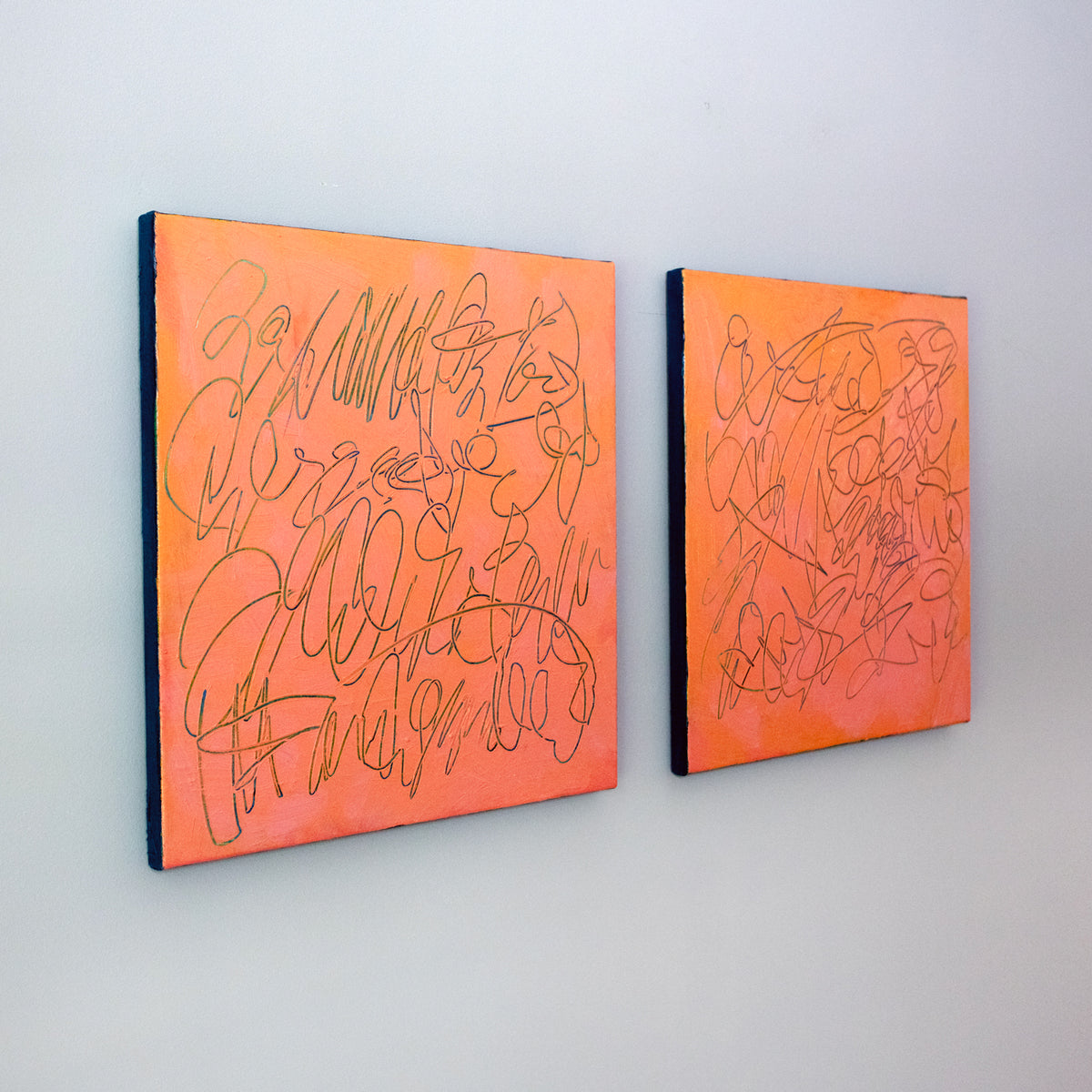 Two square abstract texted based paintings with orange and pink backgrounds and teal carved line drawings, displayed side-by-side on a light gray wall.