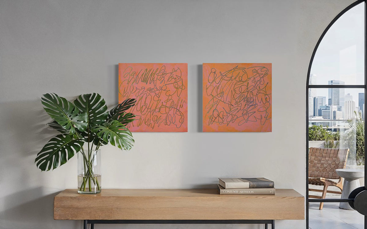 Two square abstract text based paintings with orange-pink backgrounds and thin teal scribbled lines, displayed side-by-side on a light gray wall above a wooden console table with a green plant and books. A large arched window on the right shows a city skyline and patio chair.