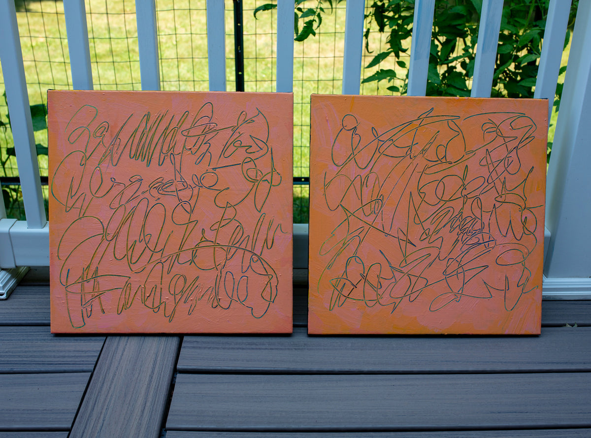 Two square abstract text based paintings with orange and peach backgrounds and teal scribbled line art, displayed side-by-side on a dark gray wooden deck in front of a white railing with green foliage in the background.