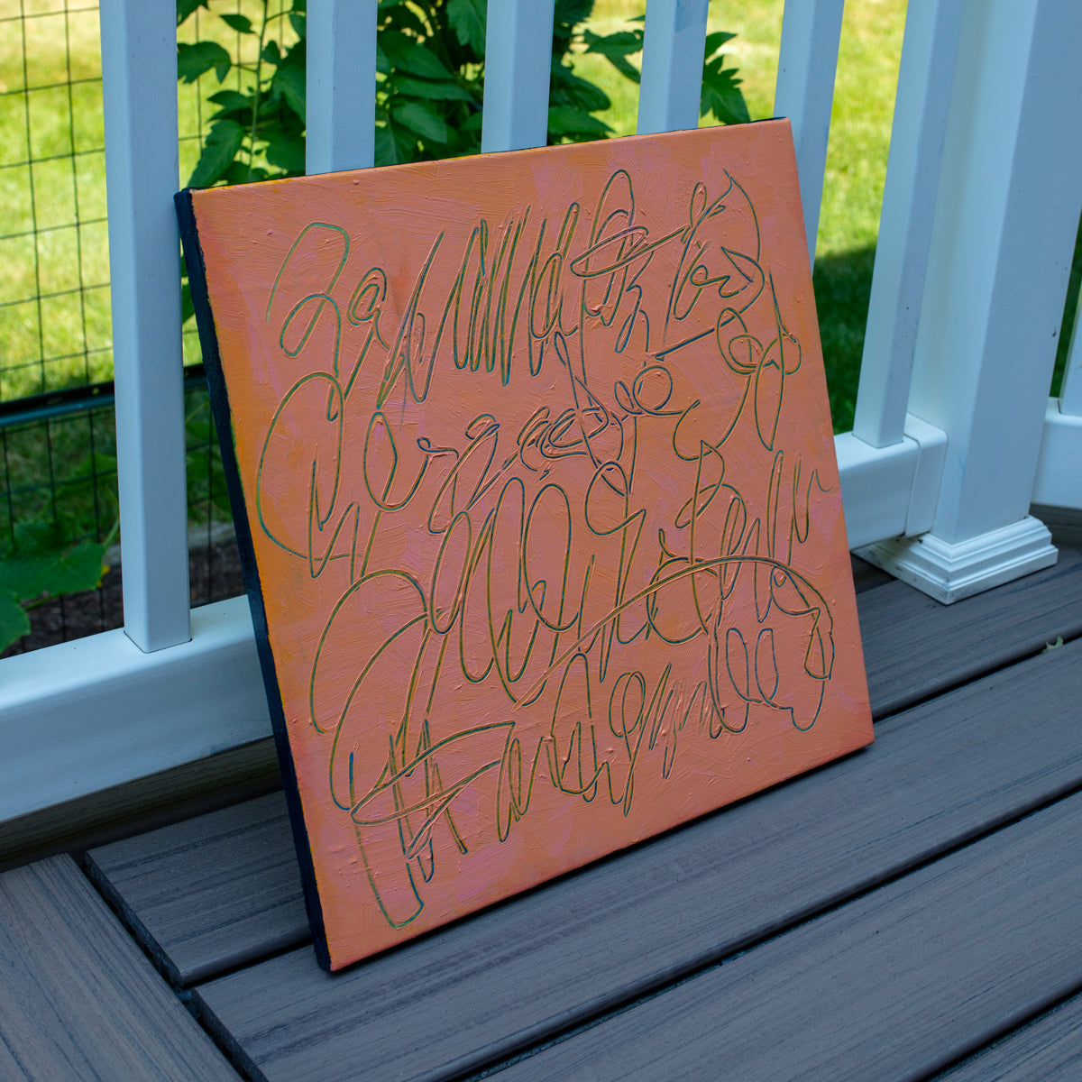 An abstract text based painting with a peach-orange background and teal carved scribbled line art, leaning against a white railing on a gray deck with green grass in the background.