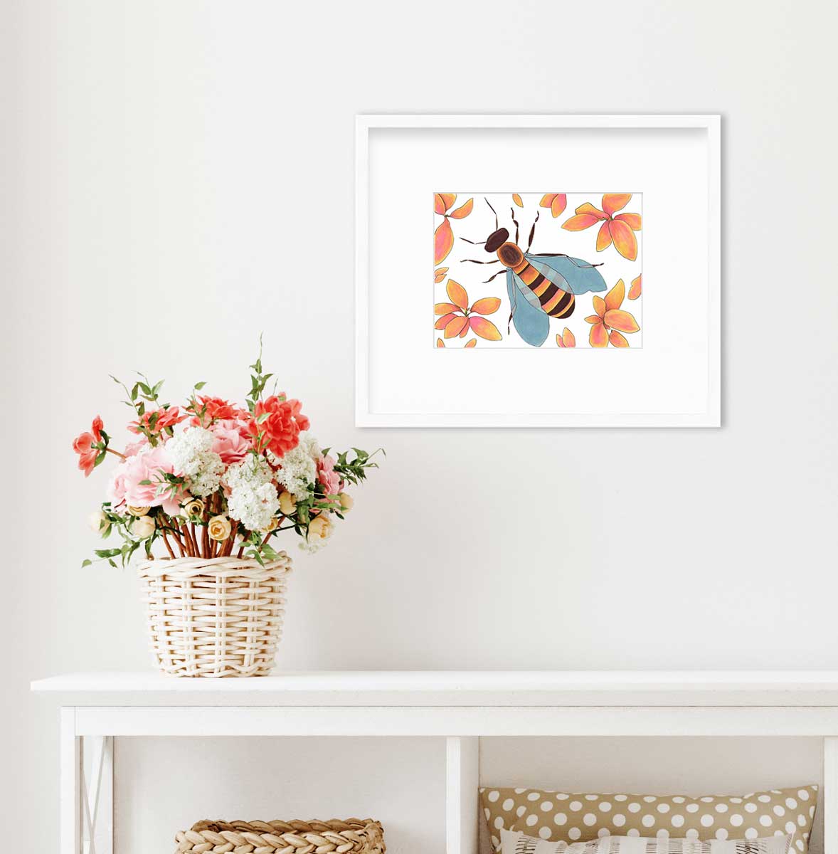 A bee surrounded by orange and pink flowers in a white frame with a mat hangs on a white wall above a white console table with storage underneath and a pink and red floral arrangement sits on top in a wicker basket.