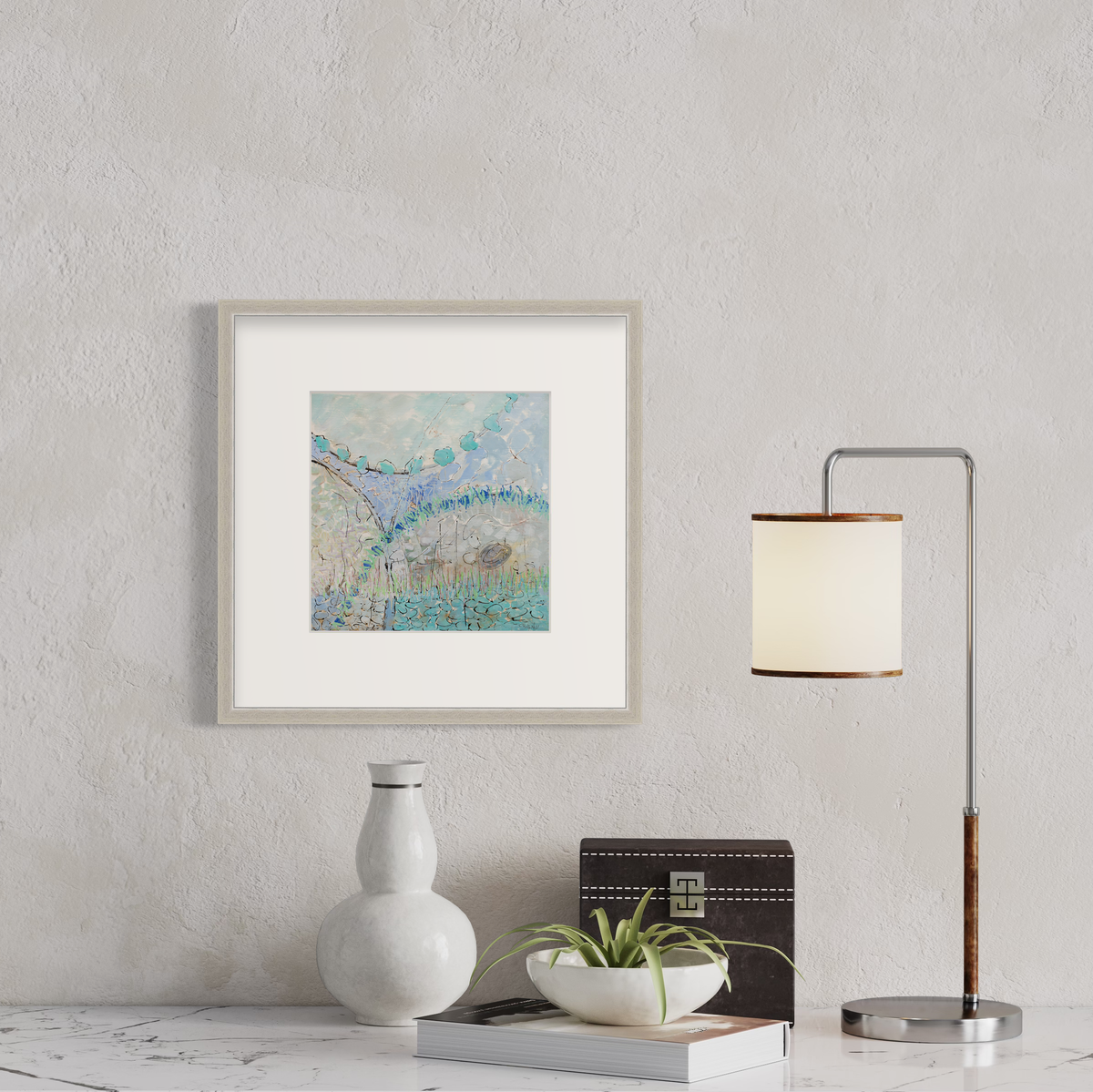 A teal, lavender, black and grey abstract print in a silver frame with a mat hangs on a white wall above a white marble surface with a small lamp and decorative accessories.