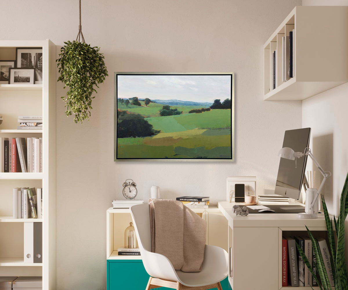 A green limited edition landscape print in a silver frame hangs on a white wall above a white workspace and next to a white bookcase. 