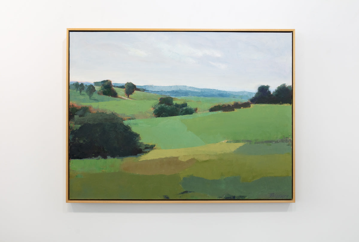 A green landscape painting framed in a light wood floater frame hangs on a white wall. 