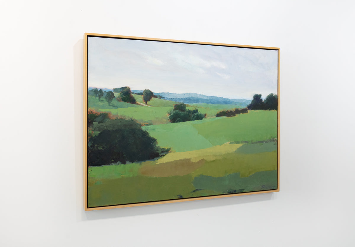 A green landscape painting framed in a light wood floater frame hangs on a white wall.