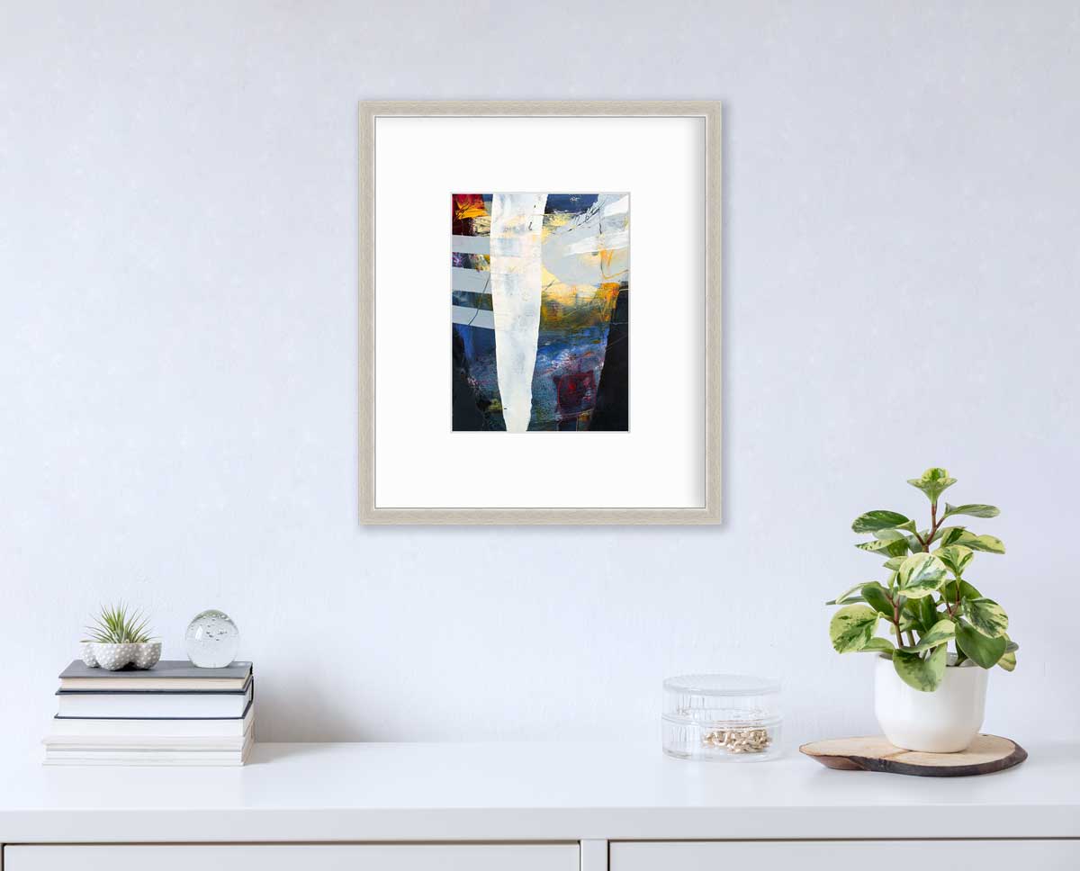 A white, yellow, red and grey abstract print in a silver frame with a mat hangs on a white wall above a white surface with a stack of books, a clear round container with gold accessories and a potted plant.
