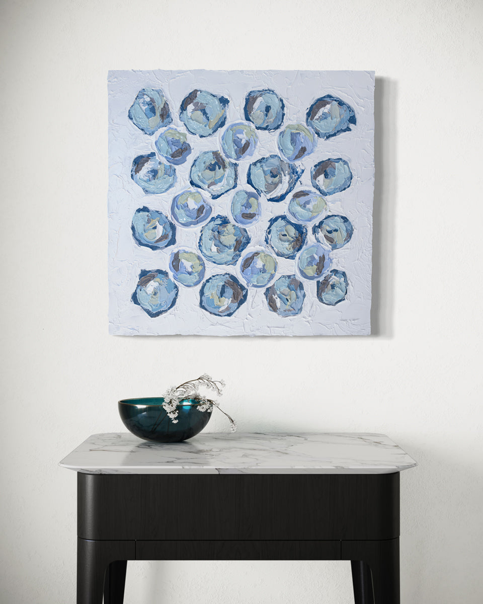 A very textured abstract painting with white background and circle type abstract flowers in blue, teal, green, plum and white hangs on a white wall over a small dark table with white marble top. A green bowl with a sprig of flowers hangs over the side.