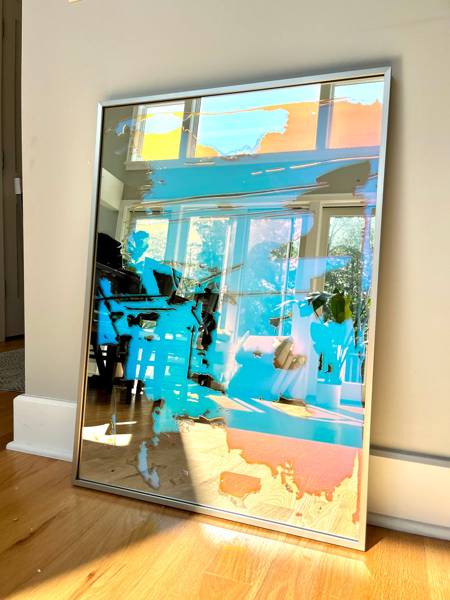 A fine art mirror rests on a wood floor against a light wall. 