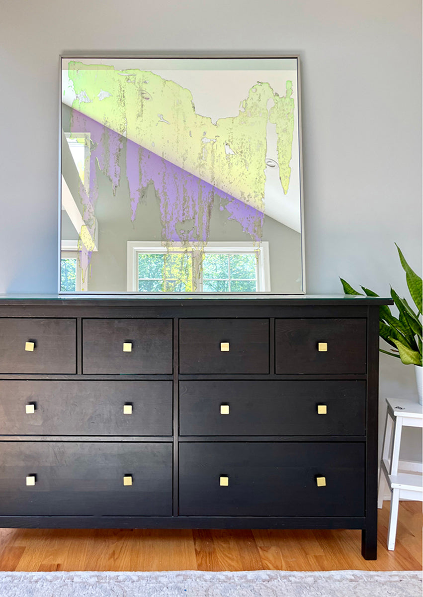 An original fine art mirror by Alina B rests on a wood dresser next to a potted plant on a white stand. 