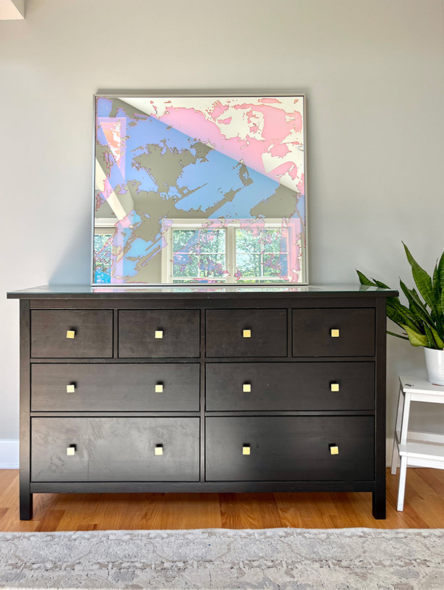 A fine art mirror rests on a wood dresser and a potted plant on a white stand. 