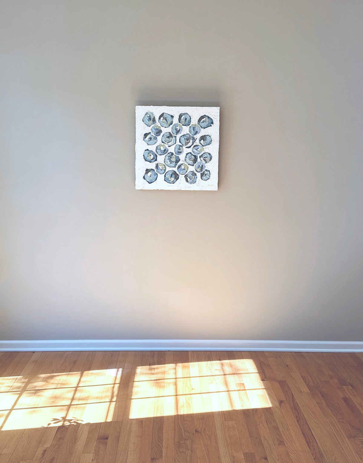 A very textured abstract painting with white background and circle type abstract flowers in blue, teal, green, plum and white hangs on a grey wall.